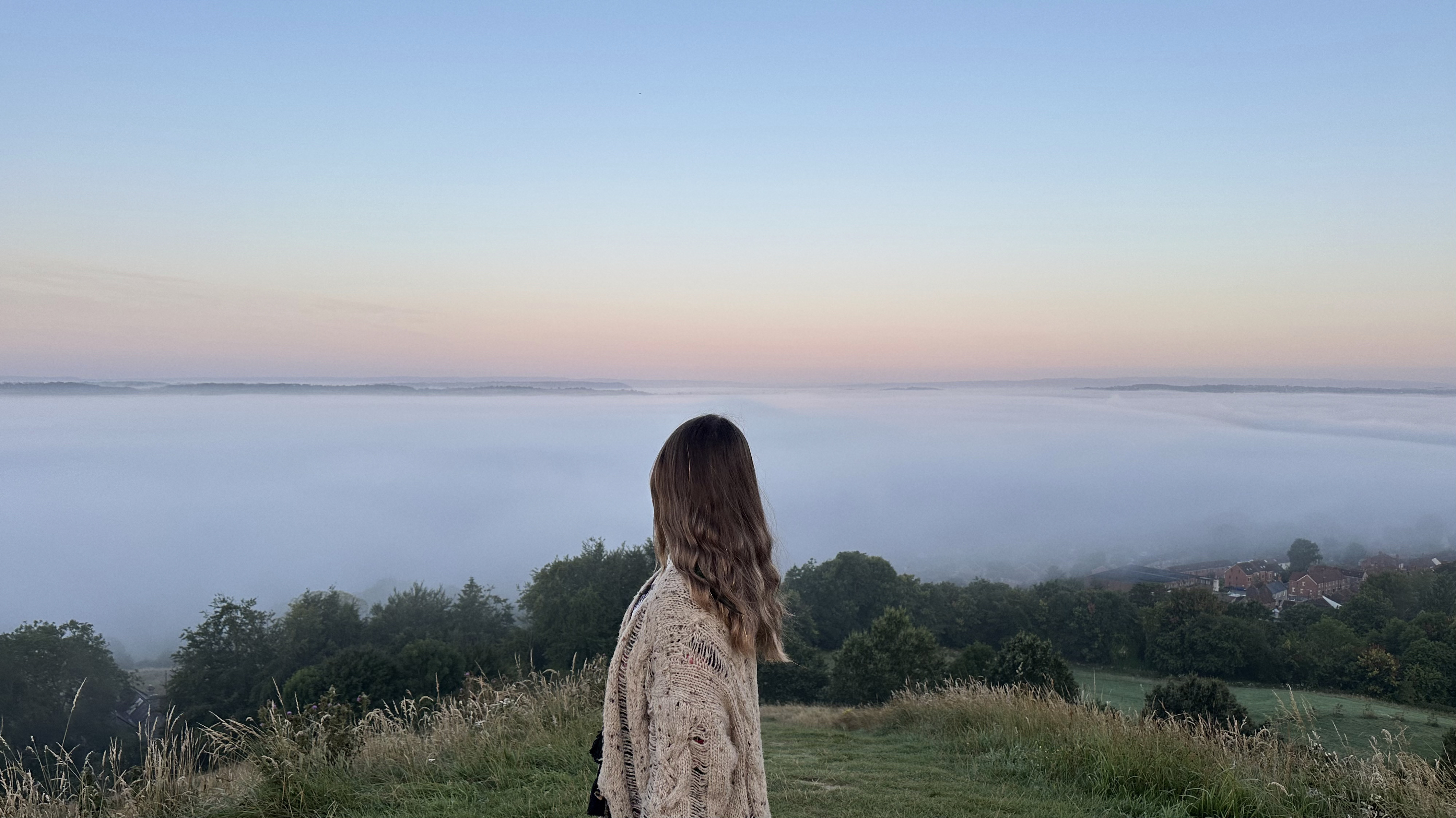Ariela de espaldas, envuelta en un cárdigan, contemplando unas colinas cubiertas por la niebla bajo un cielo suave